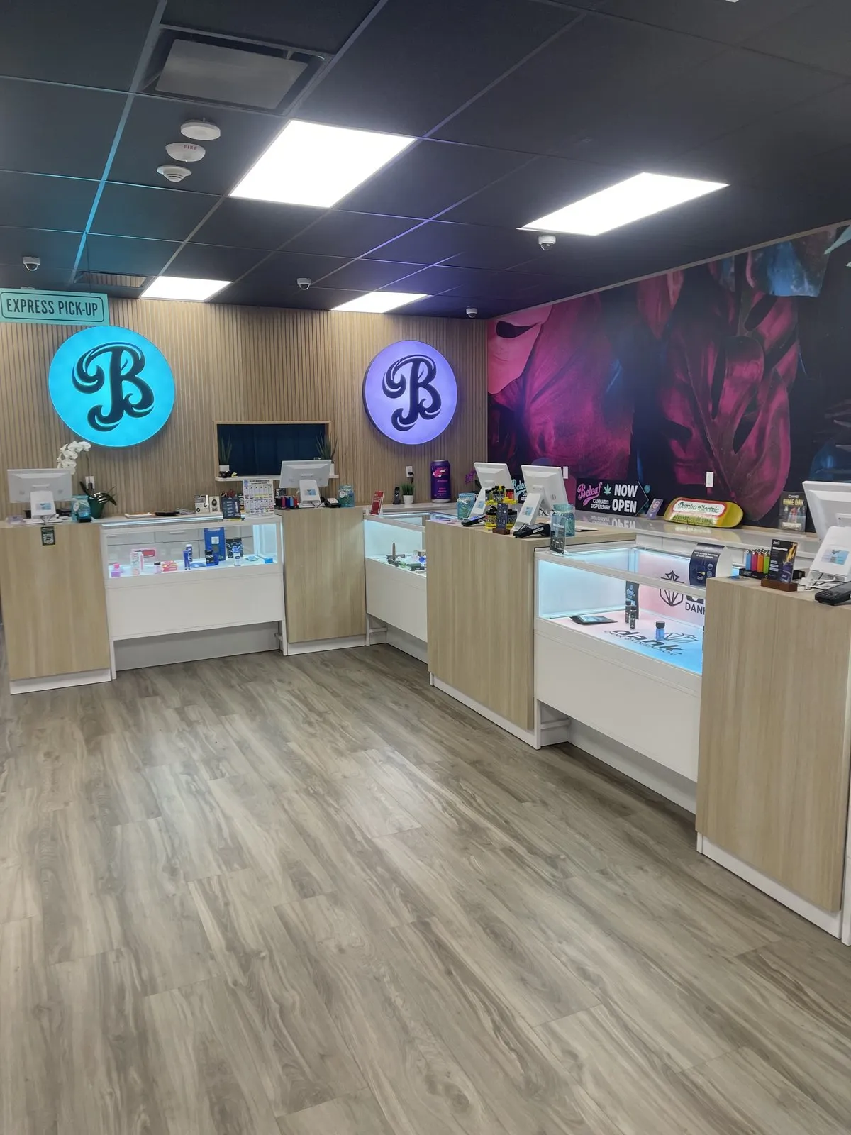 Inside Beleaf Medford - illuminated teal and purple B logos over the express pickup counter with branded product displays